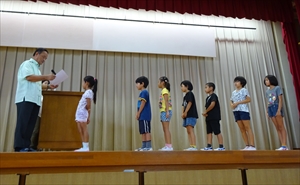平成22年度「児童・生徒の平和に関する図画・作文コンクール」表彰式で、檀上で賞状を読み上げている男性と一列に並んでいる7人の児童の写真